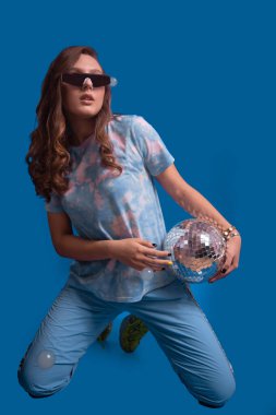 Siyah gözlüklü genç bir kadın mavi arka planda elinde bir disko topu tutuyor.