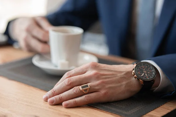 a man in a suit and with a watch in his arms drinks coffee from a white glass