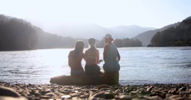 Kız arkadaşlar deniz kenarında bir kütüğe oturup dağlara bakarlar.