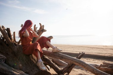 Pembe saçlı kadın sahildeki tahta kütüklere oturur ve batan güneşin tadını çıkarır.