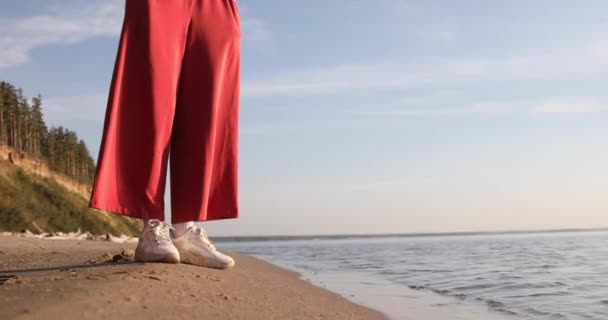 赤いパンツと白いスニーカーを着た女性が海岸に立って足で水に触れ — ストック動画