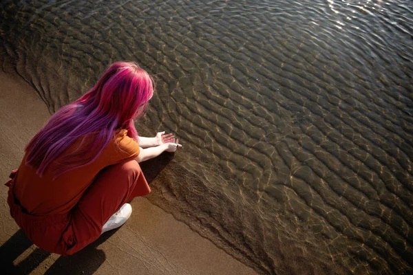 a woman with pink hair and orange clothes picks up water from the sea in her palms