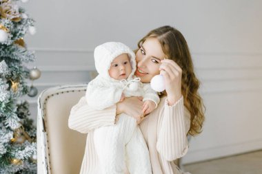 Noel ağacının arkasında küçük bir çocuğu olan mutlu bir anne. Yeni yıl kutlaması. Noel ve aile kavramı. Evdeki parlak bir odada bebeğiyle oynayan mutlu bir annenin portresi..