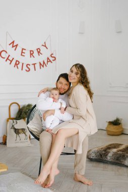 Genç ebeveynler bebeklerini kucağına alıp Noel ve yeni yıl için dekore edilmiş bir yatak odasının arka planında ona gülümsüyorlar.