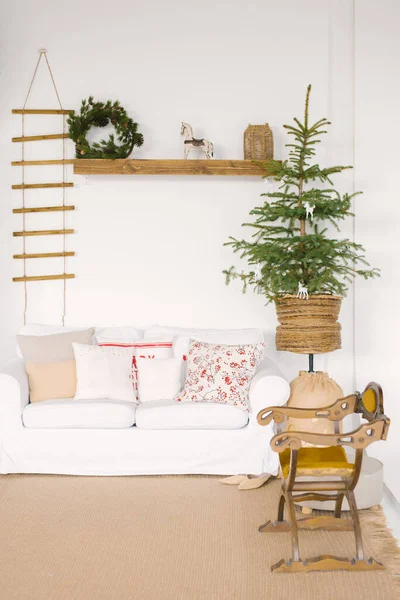 Stylish Scandinavian decor in the living room for Christmas or New Year: a white sofa with pillows, a Christmas tree, a wooden rocking chair, Souvenirs and a Christmas wreath