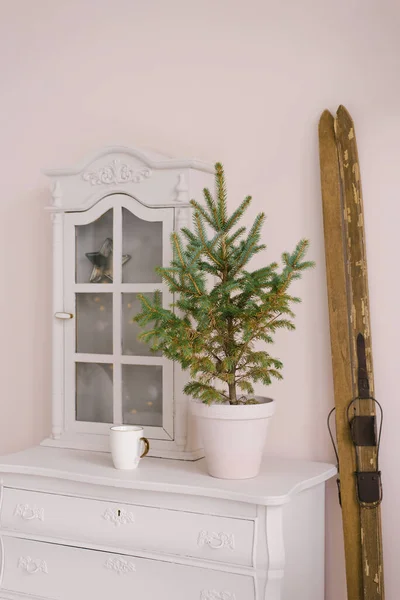 A Christmas tree in a ceramic pot stands on a white dresser in the living room, decorated for Christmas or New Year. Retro wooden skis stand near the wall