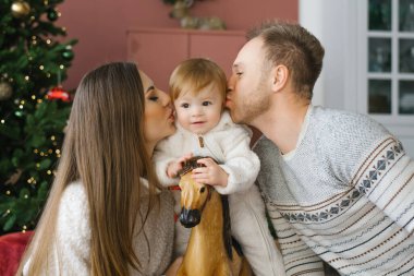 Bir yaşında küçük bir çocuk Noel ağacının yanında tahta bir atın üzerinde oturuyor. Ailesi onun yanında oturuyor ve onu yanaklarından öpüyor. Mutlu aile fotoğrafı.