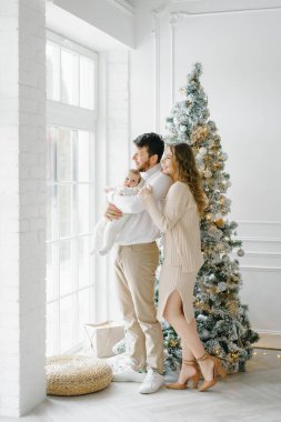 Güzel, mutlu bir aile ve küçük bir çocuk. Pencerenin yanında duruyorlar ve Noel ağacı ve birbirlerine gülümsüyorlar.