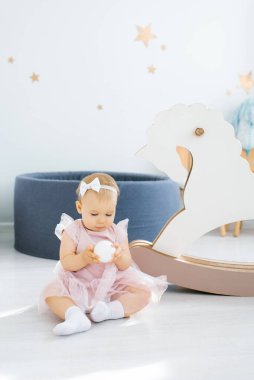 Pembe elbiseli şirin mavi gözlü bir bebek elinde küçük beyaz bir top tutuyor ve çocuk odasında yere oturuyor.
