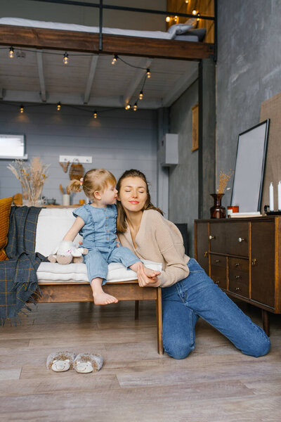 Two-year-old daughter kisses her mother on the cheek, sitting on the couch