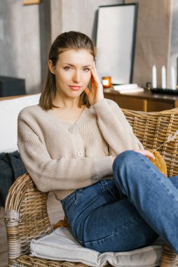 Kotlu ve kazaklı güzel bir kadın şöminenin yanındaki hasır sandalyeye oturur ve elini başına koyar.