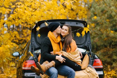 Mutlu bir çift bir arabanın bagajında oturup sonbahar manzarasının tadını çıkarır. Seyahat ve hafta sonu tatili kavramı.