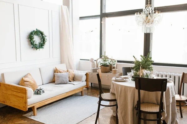 A dining table with wooden chairs, decorated and served for Christmas dinner against the background of a large window in the house, a sofa with pillows in the living room