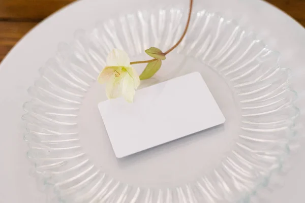 A white blank mock-up of a guest card on a glass plate and a white delicate flower . Serving a festive banquet wedding or birthday party