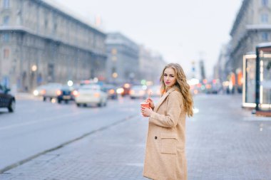 Paltolu güzel bir kız sokakta duruyor ve elinde elinde kırmızı beyaz bir bardak ve elinde bir tüp kahve var.