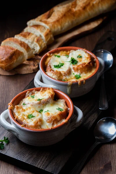 French onion soup in soup crocks with a bread baguette in behind, against a dark background.