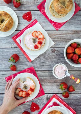 Çilek krem peyniri ve çilek dilimleriyle kaplı bir simit tutan bir elin yukarıdan görünüşü.