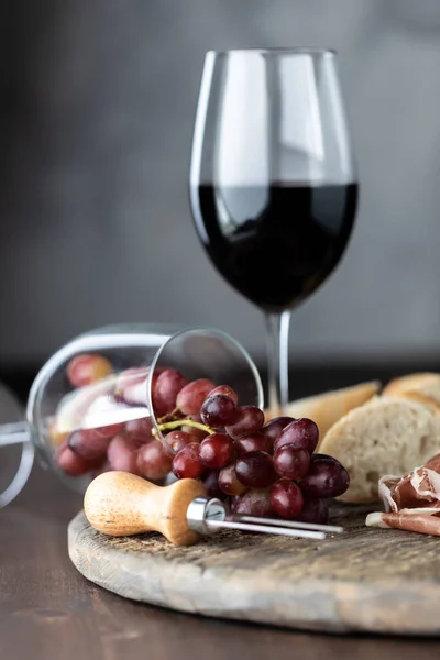 Um copo de vinho tinto com outro copo de vinho derrubado com uvas vermelhas derramando em um tabuleiro rústico. — Fotografia de Stock