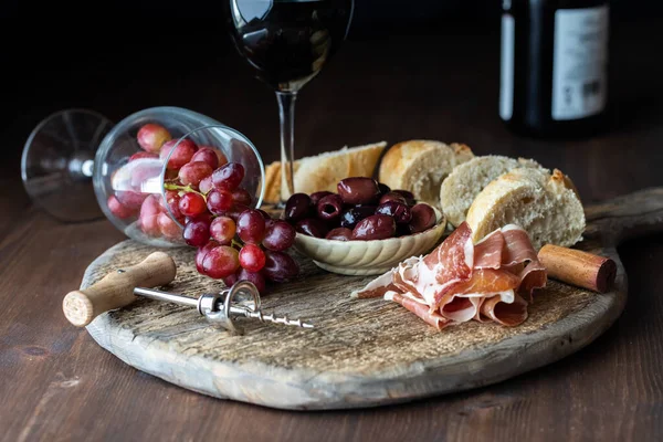 Deska Charcuterie z oliwkami, szynką i chlebem oraz szklanka wina wypełniona czerwonymi winogronami. — Zdjęcie stockowe