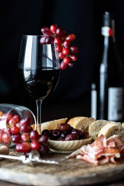 Um copo de vinho com uvas vermelhas penduradas sobre a borda contra um fundo escuro em uma placa de charcutaria rústica. — Fotografia de Stock