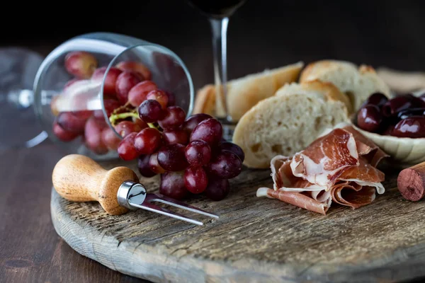 Przewrócony nad kieliszkiem wina z czerwonymi winogronami wylewającymi się na rustykalną deskę otoczoną chlebem, oliwkami i prosciutto. — Zdjęcie stockowe