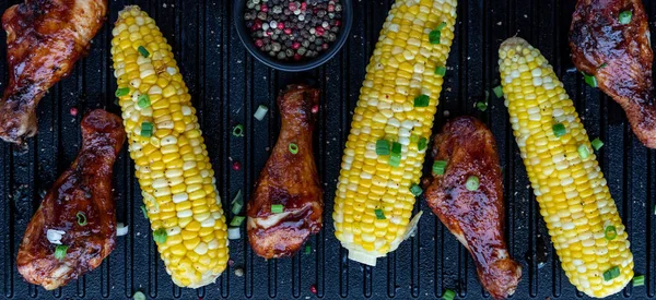 Nahaufnahme von oben auf eine Reihe von Grill-Hühnerkeulen und Mais auf dem Kolben auf einer gusseisernen Grillpfanne. — Stockfoto