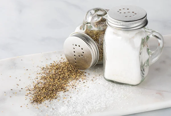 Close up de sal e pimenta shakers com sal e pimenta derramado na frente e a pimenta tombada. — Fotografia de Stock