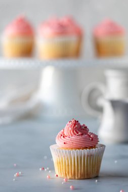 A close up of a single vanilla cupcake with rose frosting and some in soft focus in behind.