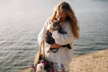 Sarışın bir kadın, yürüyüşte İskoç bir kediyi kucaklıyor.