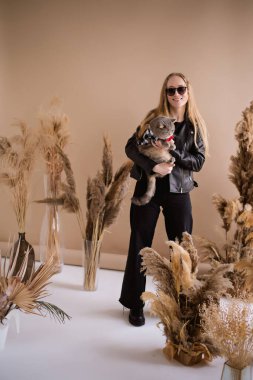 Deri ceketli, güneş gözlüklü, şık bir genç kadın, elinde neşeli bir fiyonk takan bir kedi tutuyor. Dekoratif otların arasında, bej bir arka plana karşı gülümsüyor ve modern bir cazibe yayıyor..