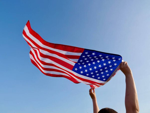 A pair of raised hands holds the United States flag, proudly waving in the air against a clear blue sky. Symbolizes freedom, patriotism, and national pride
