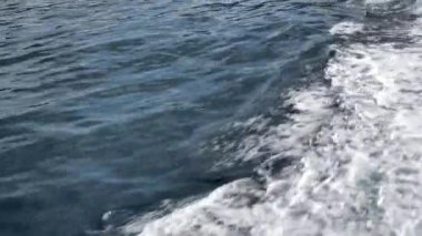 Hareket eden bir teknenin yarattığı dalgalar ve köpüklü dalgalarla deniz suyu yüzeyinin yakın görüntüsü. Dinamik su dokusu, derin mavi okyanus yansımaları, seyahat konsepti, deniz arka planı
