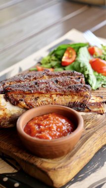 Köy tahtasında servis edilen leziz ızgara pirzolaların dikey yakın çekimi. Tabağa bir kase zengin sos ve taze salata eşlik eder ve ağız sulandırıcı bir yemek oluşturur..