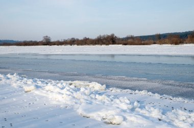 Ufuk boyunca kısmen donmuş bir nehir, karla kaplı kıyılar ve çıplak ağaçların yer aldığı sakin bir kış sahnesi. Sakin mavi gökyüzü, doğa, seyahat ve yaşam tarzı projeleri için uygun kış havasını tamamlıyor..