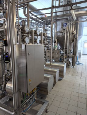 Temiz, iyi ışıklandırılmış bir fabrikada paslanmaz çelik borular, göstergeler ve kontrol panelleri bulunan yüksek teknolojili bir endüstriyel ortam. Karmaşık sistemler ve ekipmanlar hassasiyet iletir