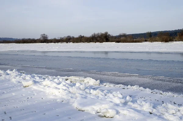 Ufuk boyunca kısmen donmuş bir nehir, karla kaplı kıyılar ve çıplak ağaçların yer aldığı sakin bir kış sahnesi. Sakin mavi gökyüzü, doğa, seyahat ve yaşam tarzı projeleri için uygun kış havasını tamamlıyor..