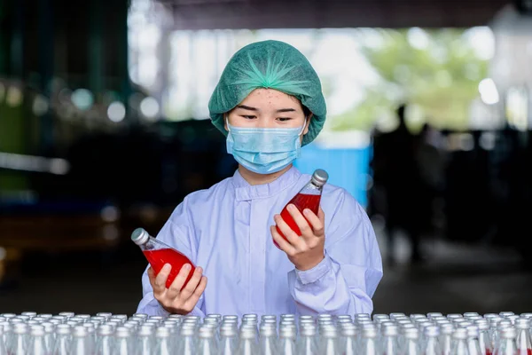 Worker of science in bottle beverage factory wearing safety  hat and face mask working to check quality of drink Basil seed produce on conveyer belt before distribution to market business