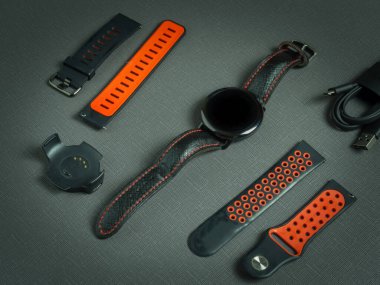 Bilek yuvarlak spor akıllı saat, kırmızı iki silikon kayış, koyu arkaplanda şarj aleti ve kadranda zaman.