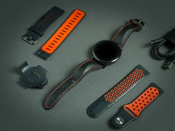 Bilek yuvarlak spor akıllı saat, kırmızı iki silikon kayış, koyu arkaplanda şarj aleti ve kadranda zaman.