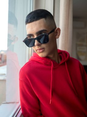 Sönük saçlı, koyu güneş gözlüğü takan ve canlı kırmızı kapüşonlu şık bir genç, pencerenin yanında duruyor. O, düşünceli bir şekilde başka yere bakıyor. Doğal ışık, onun profilini ve gelişigüzel kentsel tarzını vurgulayan yumuşak bir atmosfer yaratır.