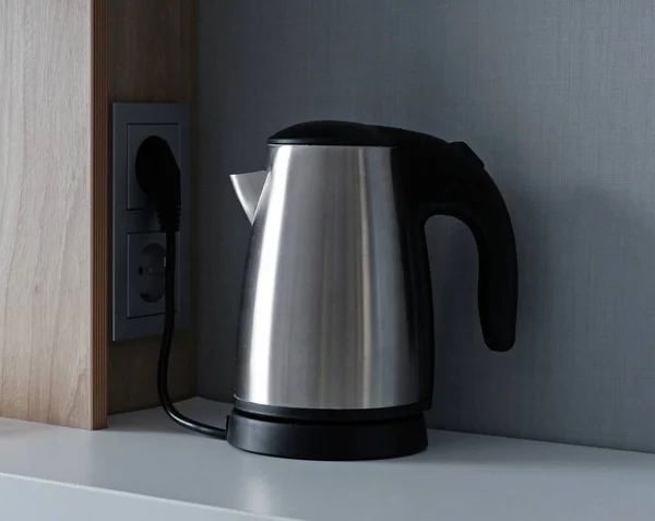 Silver metal electric kettle for boiling water and making tea or coffee on a table. Household kitchen appliances for makes hot drinks. Stainless steel electric kettle is plugged into a power outlet.