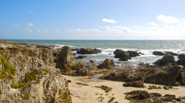 Kıyı şeridine güçlü dalgaların çarptığı engebeli kayalık sahilin panoramik manzarası. Sert bitki örtüsüyle sarp kayalıklar, engin çalkantılı deniz ve kumlu bölgeleri tamamlıyor..