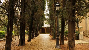 Uzun selvi ağaçları ile kaplı bahçe sokağı - Akdeniz selvisi, klasik siyah sokak lambası, yeşil alanların ortasında kemerli kireç şapeli girişine giden kaldırım taşı yolu..