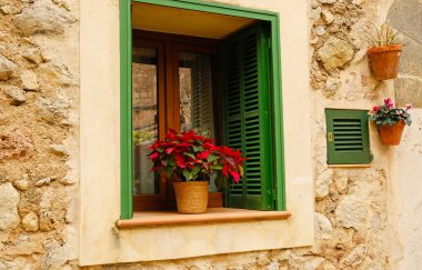 Yeşil ahşap panjurları ve kırmızı poinsettia kabı olan taşralı taş bir pencere. Valldemossa, Mallorca 'da geleneksel Akdeniz ön cephe dekorasyonu.
