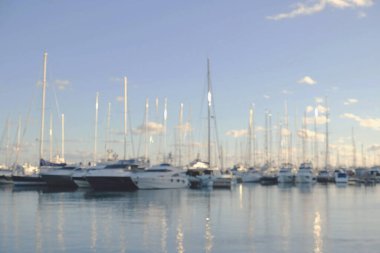 Lüks yatları ve sakin mavi sulara yansıyan yelkenli direkleri olan hafif bulanık marina. Seyahat, kiralama, deniz kenarı gayrimenkul ve yaşam tarzı markaları için soyut denizcilik geçmişi.