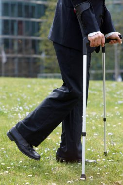 İş adamı, dizinden sakatlandıktan sonra koltuk değnekleriyle şehirde bir parkta yürüyor.