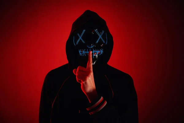 Anonymous man in a hoodie hiding face behind neon glow scary mask on red background. Horror concept