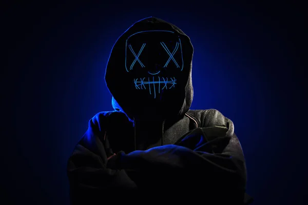 Anonymous man in a hoodie hiding face behind neon glow scary mask on dark background. Horror concept
