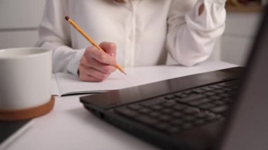 Kadın mutfaktaki iş yerinde uzaktan çalışıyor. Kadın dizüstü bilgisayarda çevrimiçi ders dinlerken not defterine not alıyor. Uzaklık eğitimi ve e-öğrenim kavramı.