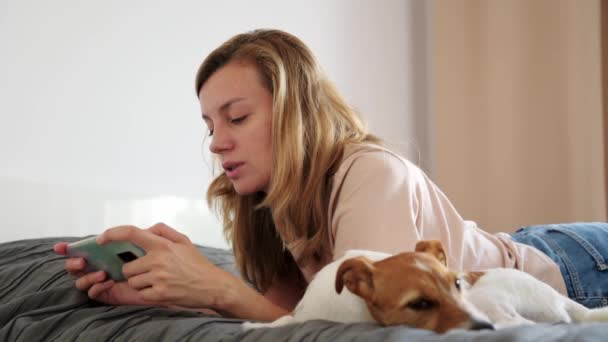 Женщина с собакой лежит на кровати и использует смартфон — стоковое видео